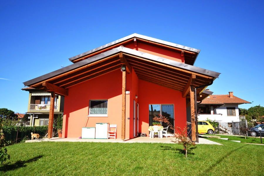 Casa rossa con tetto spiovente su un prato soleggiato, con altre case sullo sfondo.