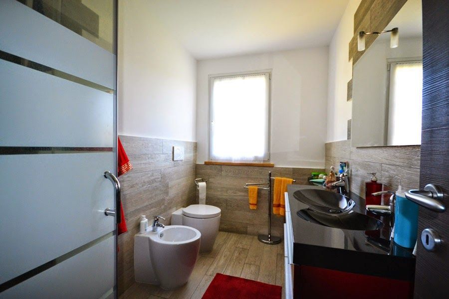 Piccolo bagno con lavandino, WC, bidet, doccia e pareti piastrellate illuminate da una finestra.