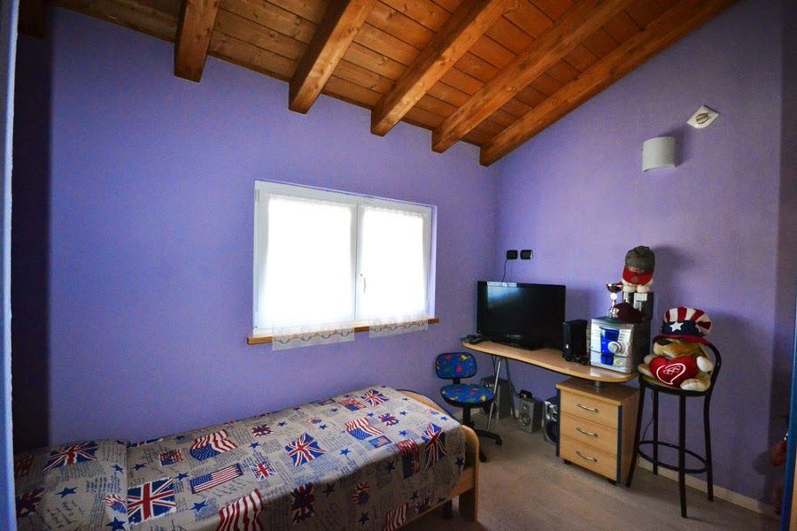 Camera da letto viola con soffitto in legno, letto, finestra e scrivania con TV e macchina del caffè.
