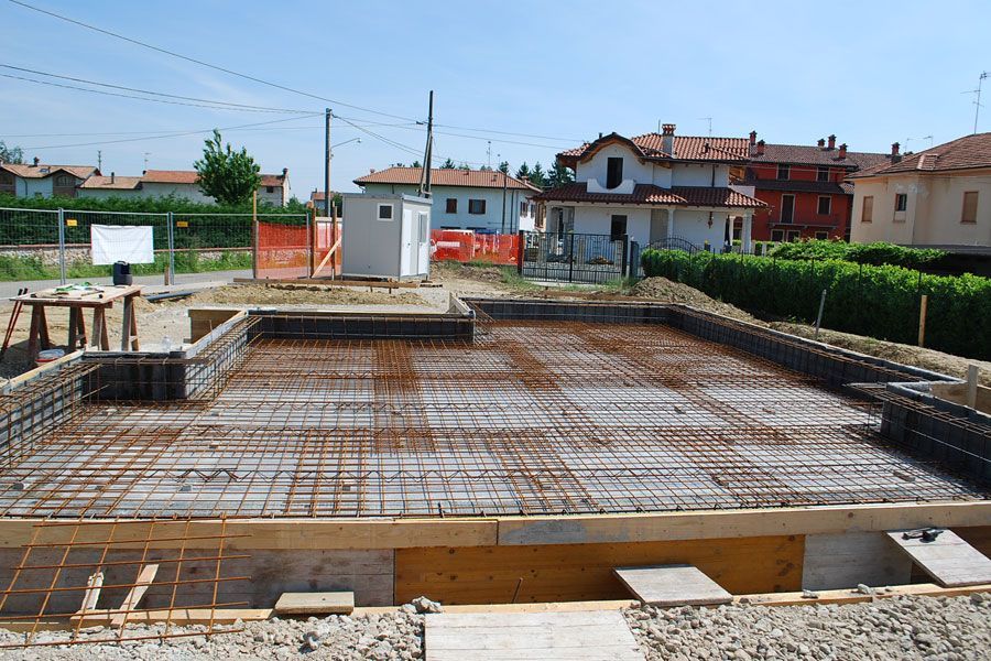 Soletta di fondazione in cemento in costruzione nel cortile di una casa, con armature in ferro e case vicine sotto un cielo azzurro.
