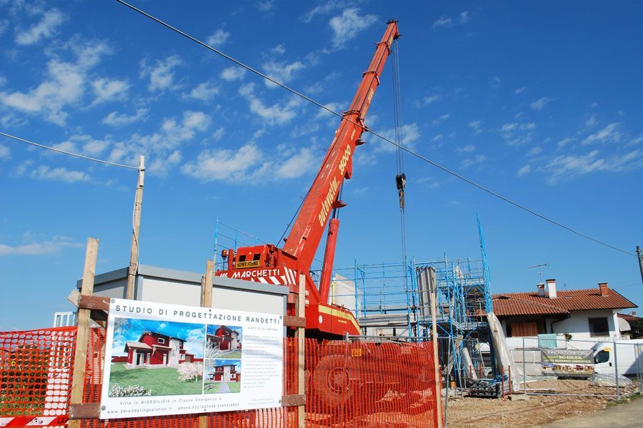 Cantiere edile con gru arancione, impalcature e un cartello recintato sotto un cielo azzurro.