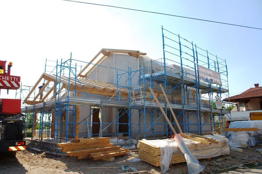 Casa in costruzione con impalcature blu e legname accatastato in una soleggiata zona residenziale.