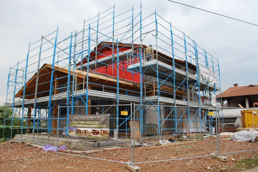 Casa in costruzione con impalcature blu e struttura del tetto rossa a vista.