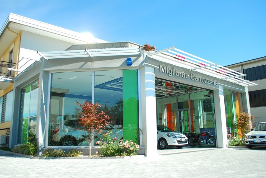Concessionaria automobilistica moderna con facciata in vetro, un'auto sportiva bianca all'interno e un cielo azzurro all'esterno.
