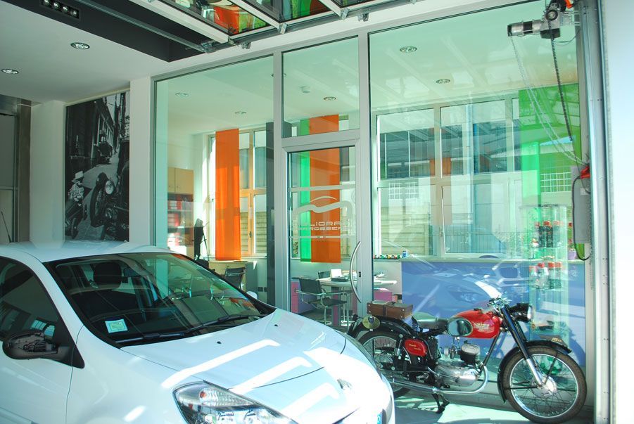 Un'auto bianca parcheggiata in uno showroom con pareti di vetro, accanto a motociclette e insegne arancioni.