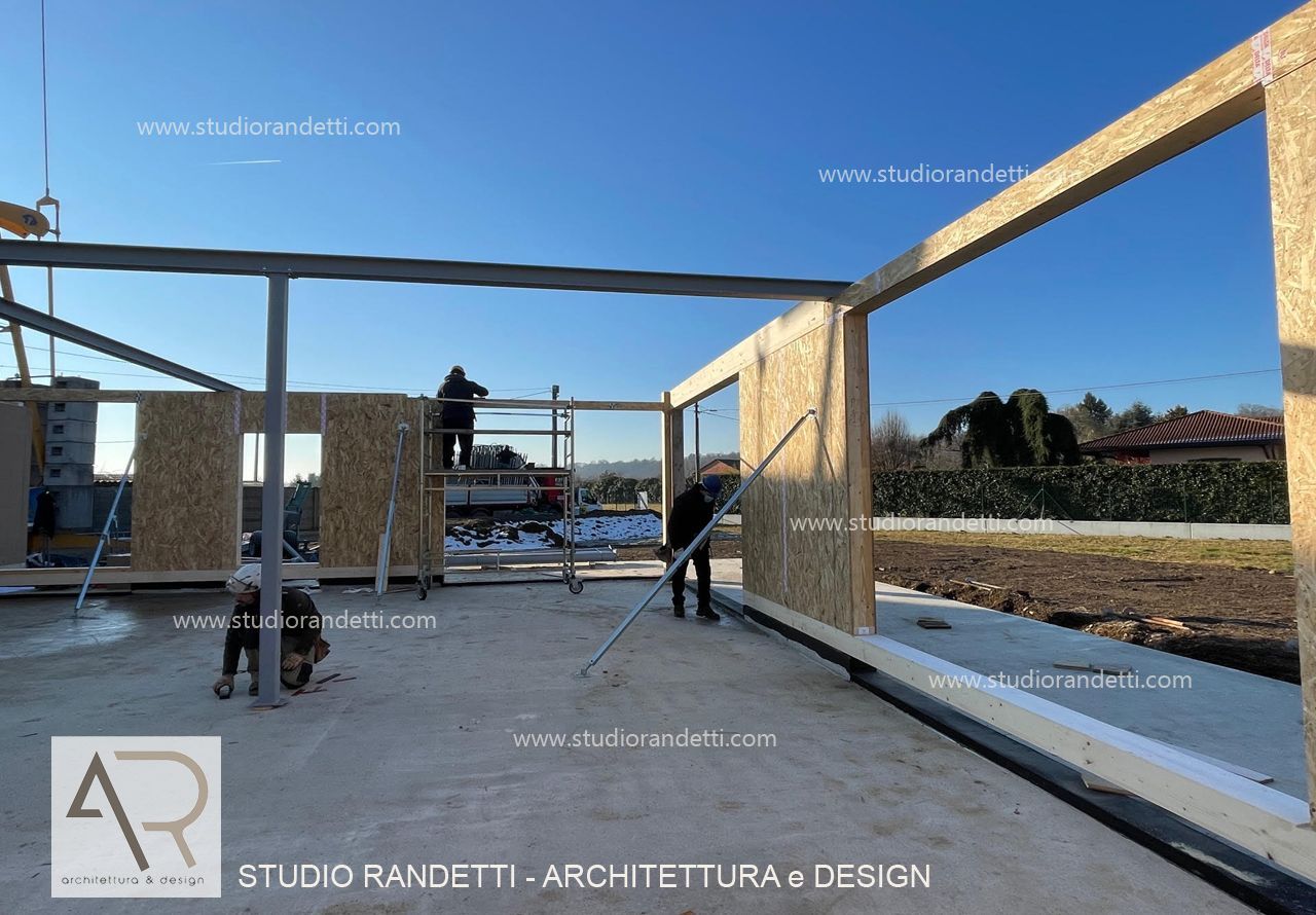 Struttura in legno parzialmente costruita sotto un cielo azzurro e limpido, con due operai intenti ad assemblare le pareti.