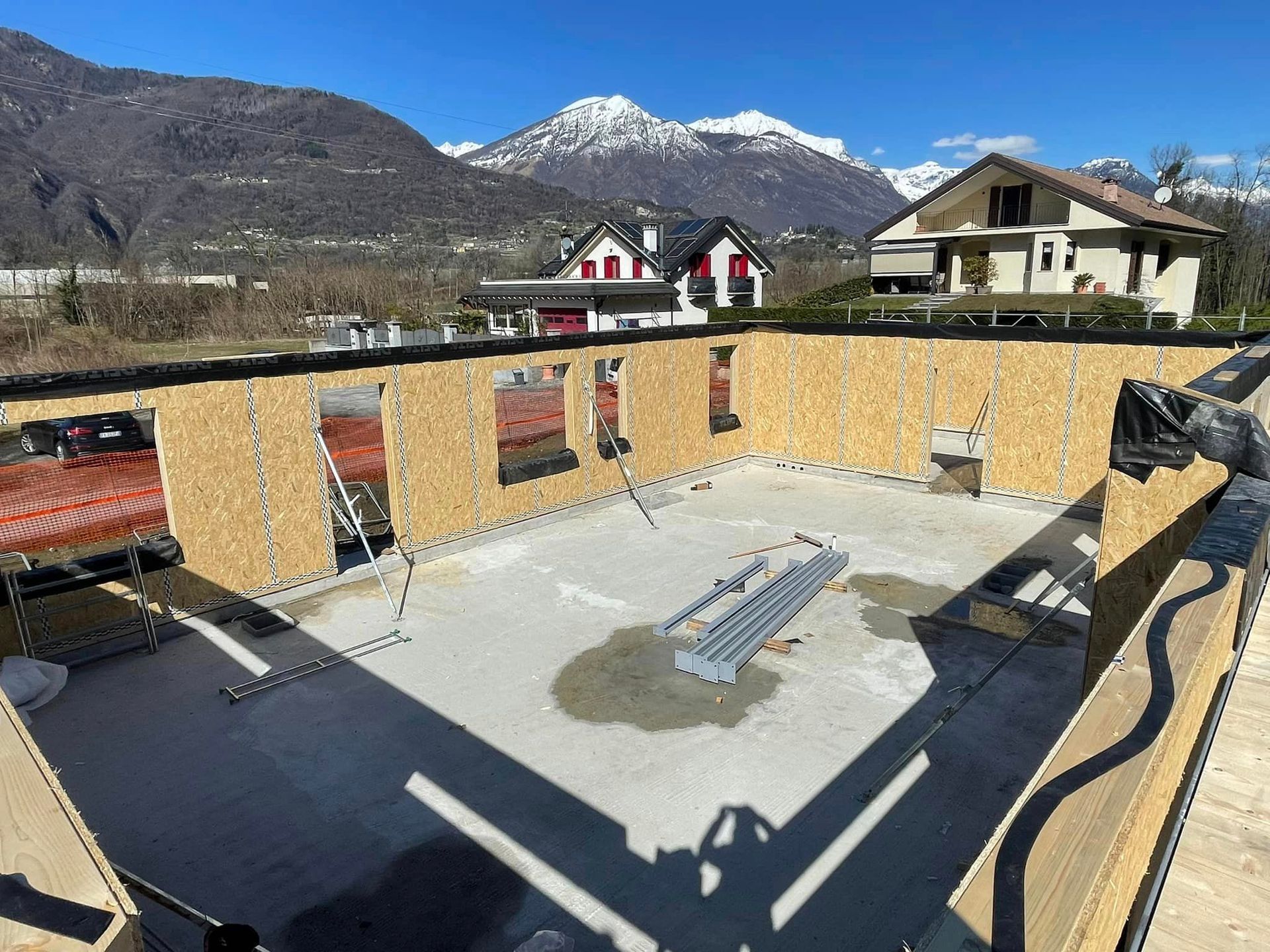 Terrazza sul tetto con panchine e tavoli in cemento, circondata da muretti bassi, con montagne e case sullo sfondo.