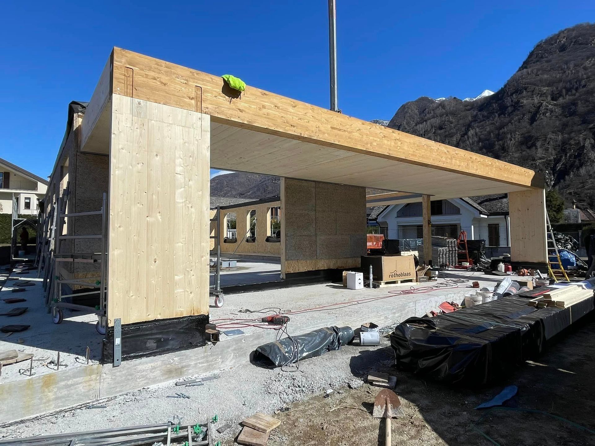 Struttura in legno parzialmente costruita in un cantiere innevato con le montagne sullo sfondo.