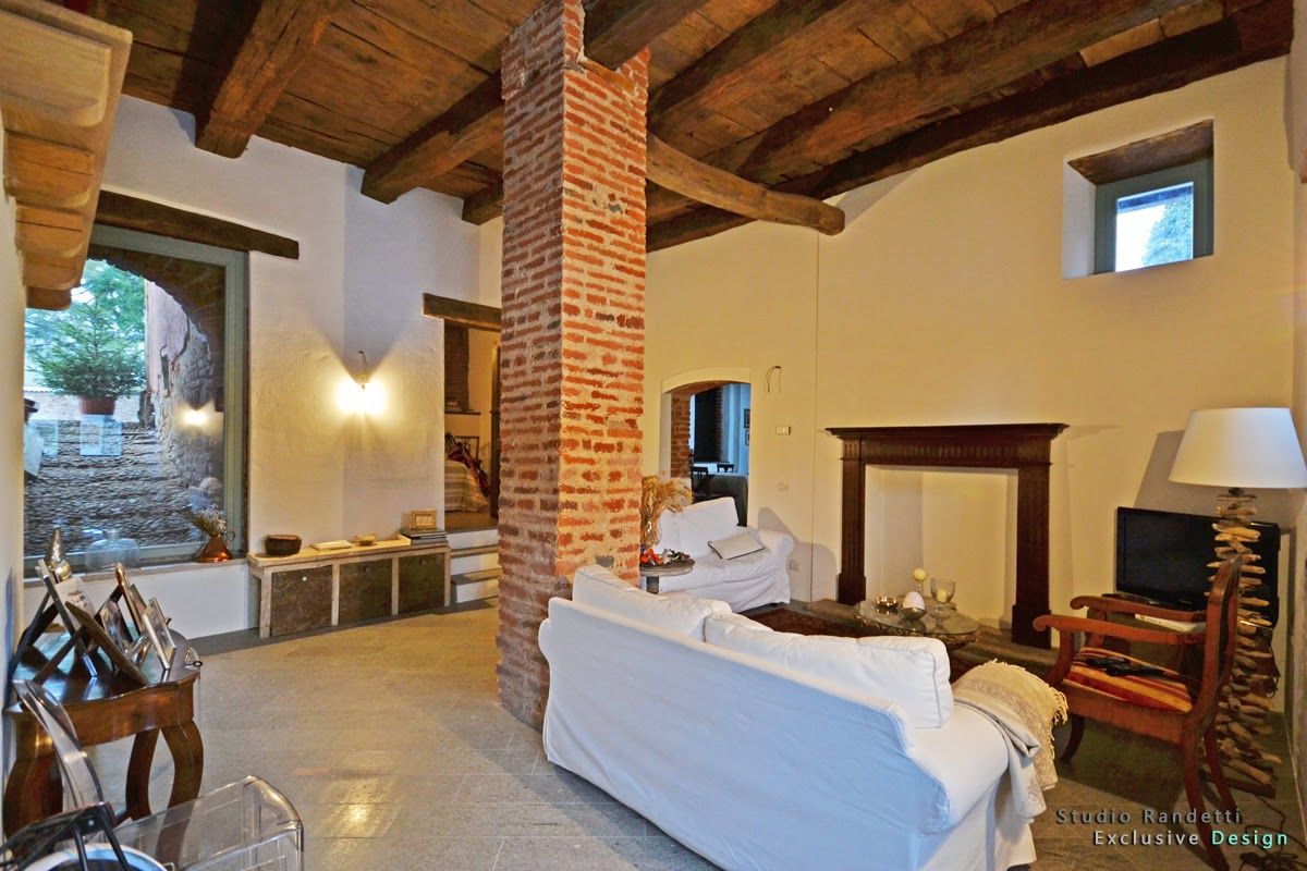 Soggiorno rustico con travi a vista, divani bianchi, camino in mattoni e porta aperta su un patio soleggiato.