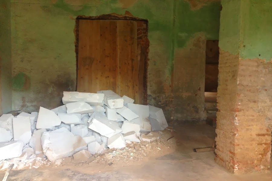 Stanza parzialmente demolita con blocchi bianchi impilati, pareti verdi, una porta di legno e pilastri di mattoni.