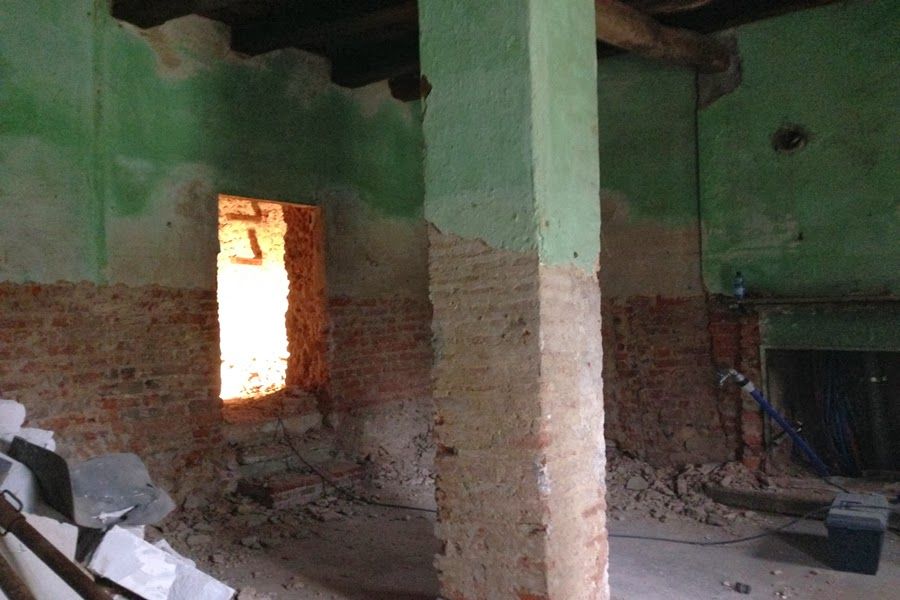 Interno di una stanza danneggiato, con muri fatiscenti, mattoni a vista e una finestra luminosa.