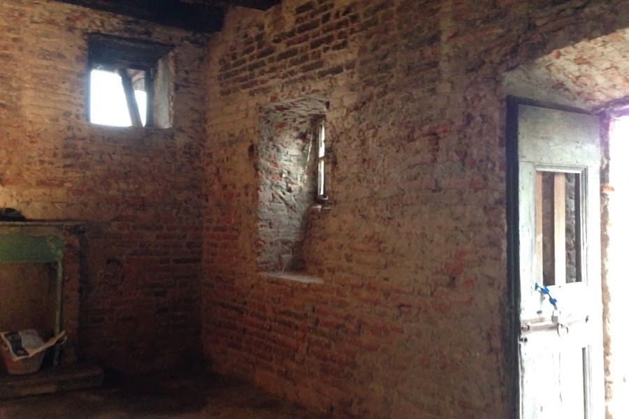 Stanza in mattoni a vista con piccole finestre e una porta bianca, illuminata dalla luce naturale.