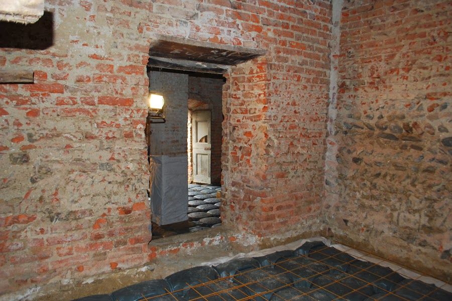 Stanza in mattoni a vista con una porta che si apre su un corridoio in pietra illuminato.