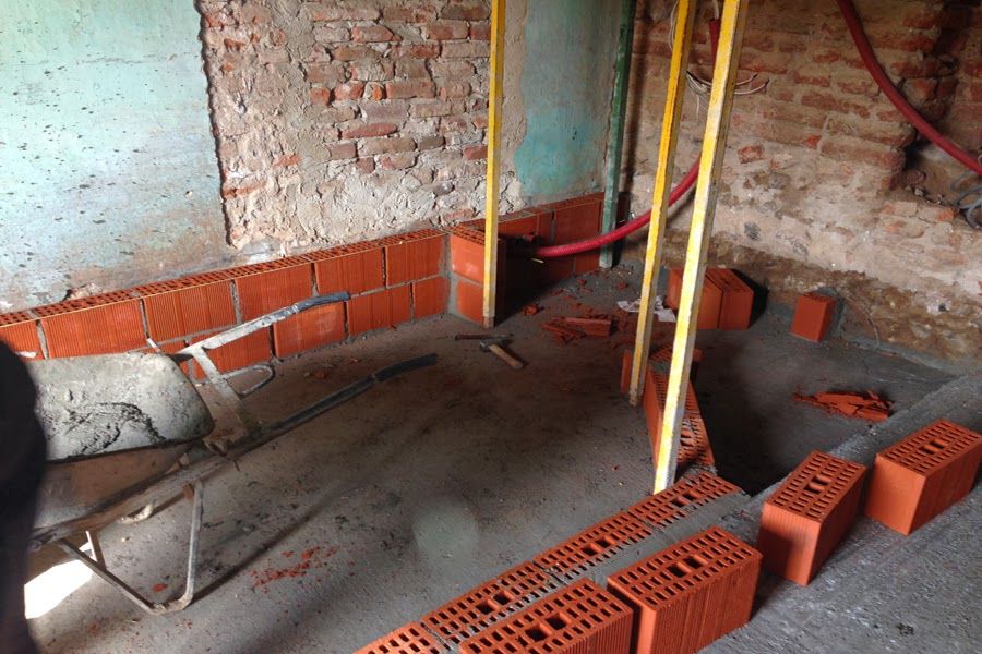 Stanza parzialmente costruita con pareti in mattoni a vista, pavimento in cemento e mattoni rossi impilati vicino a pali di sostegno gialli.