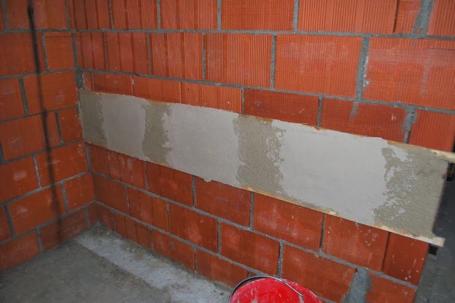 Muro di mattoni rossi parzialmente intonacato in un cantiere, con un secchio rosso sul pavimento.