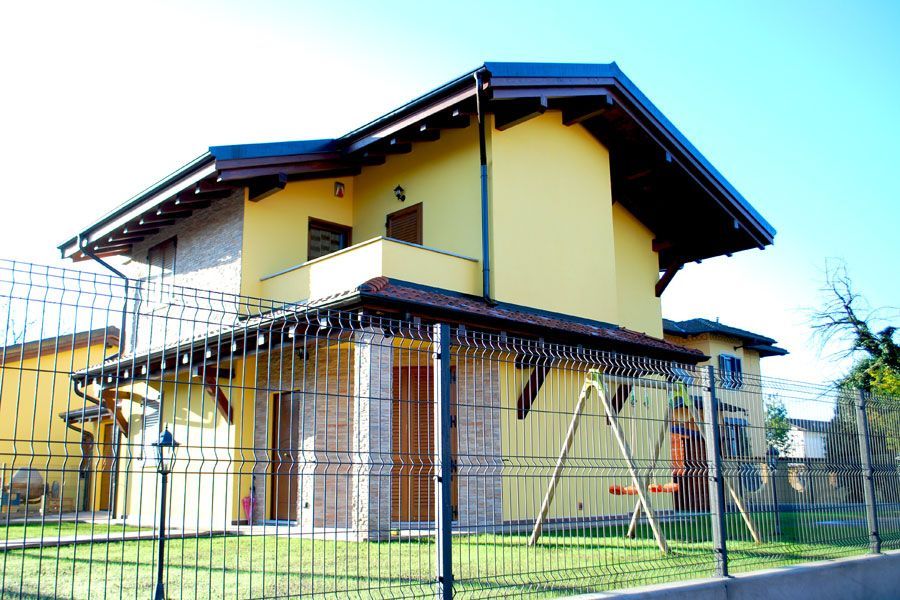 Casa moderna a due piani di colore giallo con tetto rosso e cortile recintato sotto un cielo azzurro.