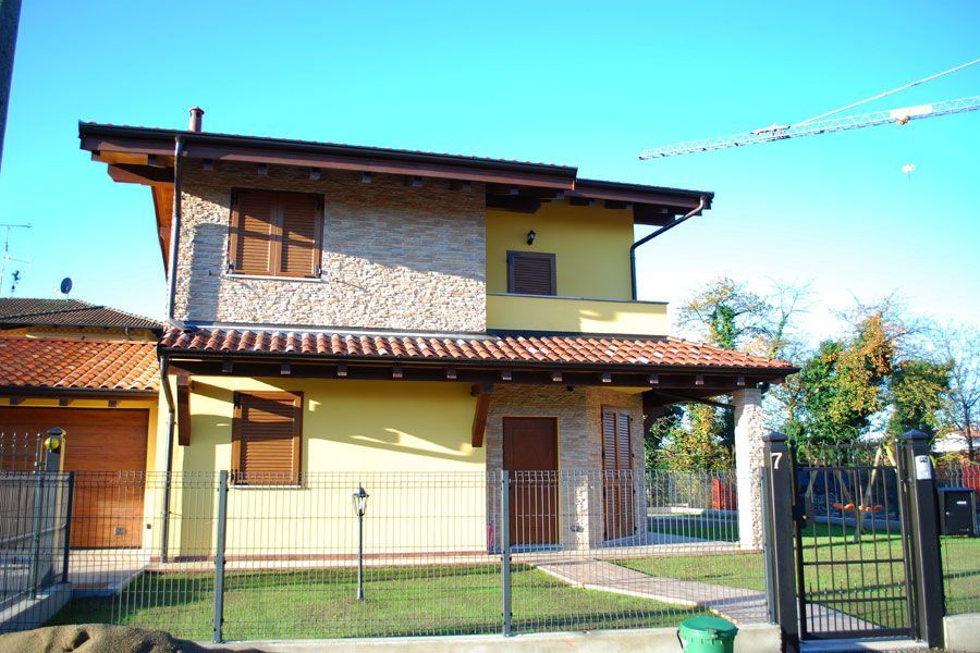 Casa a due piani con rivestimento in pietra e giallo, cortile anteriore recintato e cielo azzurro limpido.