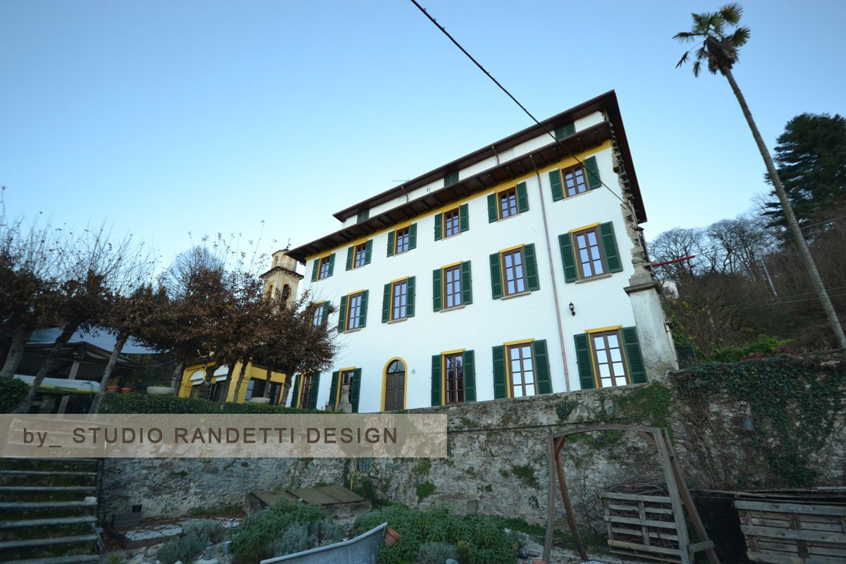Edificio bianco con persiane verdi su una collina, visto dal basso con alberi e un cielo azzurro limpido.