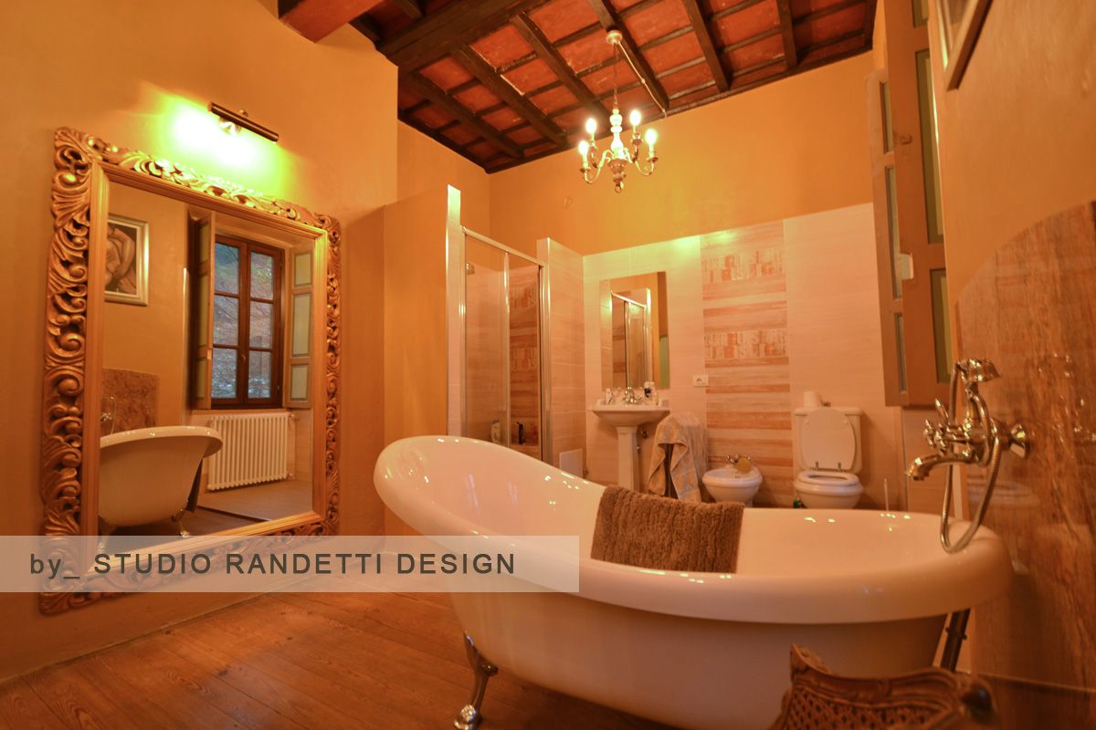 Bagno accogliente, illuminato da una luce ambrata, con vasca con piedini, doccia, mobile lavabo e soffitto con travi in legno.