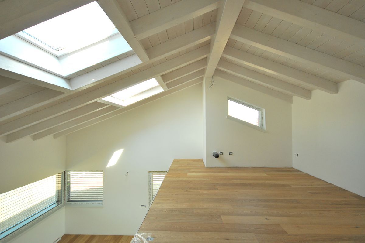 Luminoso loft mansardato con pareti bianche, travi a vista, lucernari e pavimento in legno.