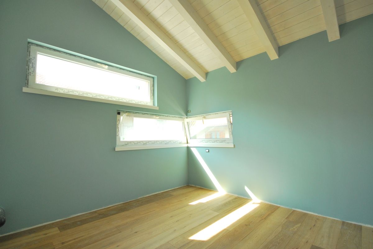 Stanza vuota con pareti color menta, soffitto inclinato in legno, finestre strette e luce del sole sul pavimento in legno.