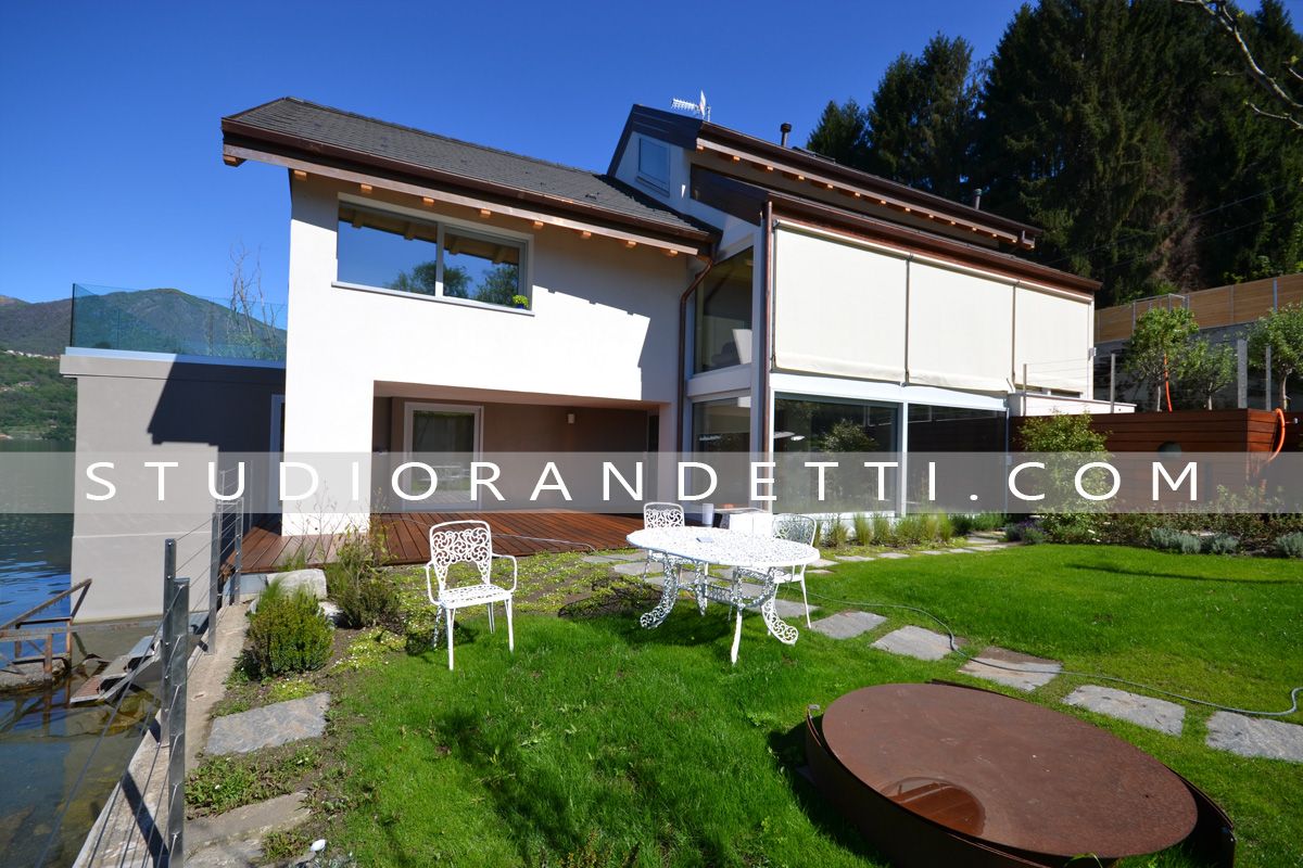 Casa moderna bianca con tetto spiovente, prato verde, mobili da giardino e un braciere rotondo sotto un cielo azzurro limpido.