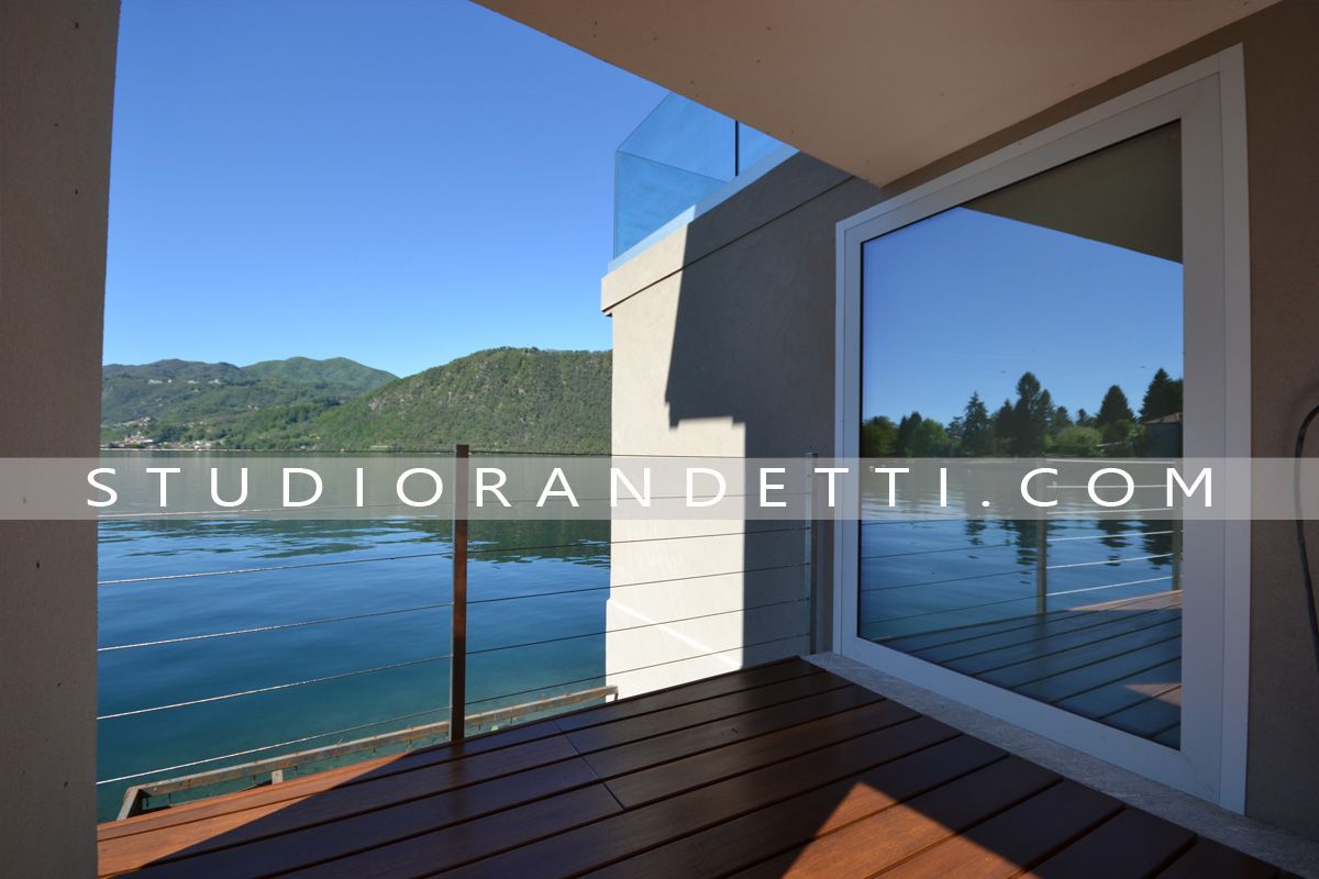 Balcone con pavimento in legno che si affaccia su un mare blu e su verdi colline attraverso un'ampia porta a vetri.