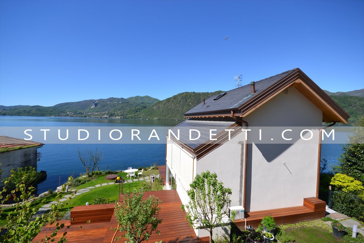 Casa bianca con tetto marrone affacciata su un lago, sulle montagne e su una terrazza con tegole rosse, immersa nella luce del sole.