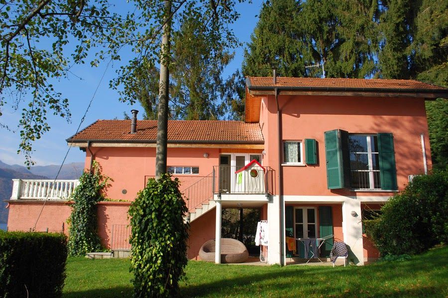Casa rosa con tetto rosso, balcone bianco e persiane verdi in un cortile soleggiato