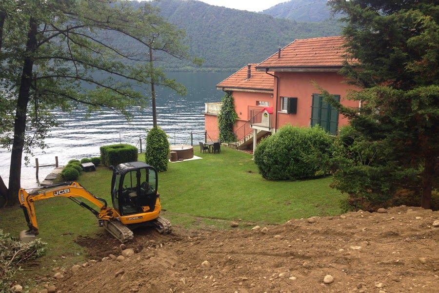 Un piccolo escavatore scava vicino a una casa in riva al lago, con le montagne sullo sfondo.