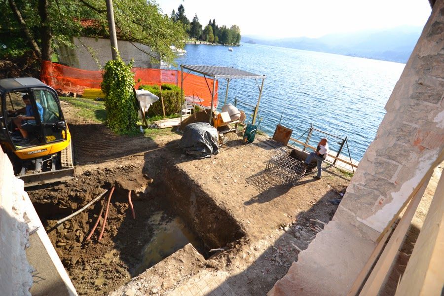 Cantiere edile in riva al lago con fossa di scavo, macchinari, operai e recinzione di sicurezza arancione.