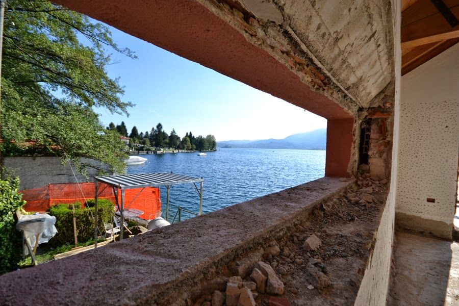 Vista sul lago attraverso una finestra rustica, con acqua azzurra, alberi in lontananza e sedie arancioni su un patio in pietra.