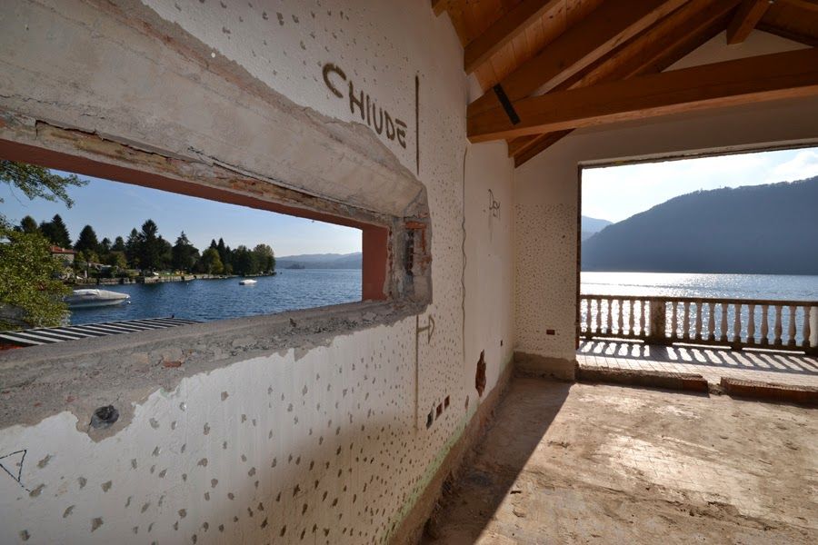 Una stanza bianca che si apre su un lago e sulle montagne, con un balcone e delle barche all'esterno.