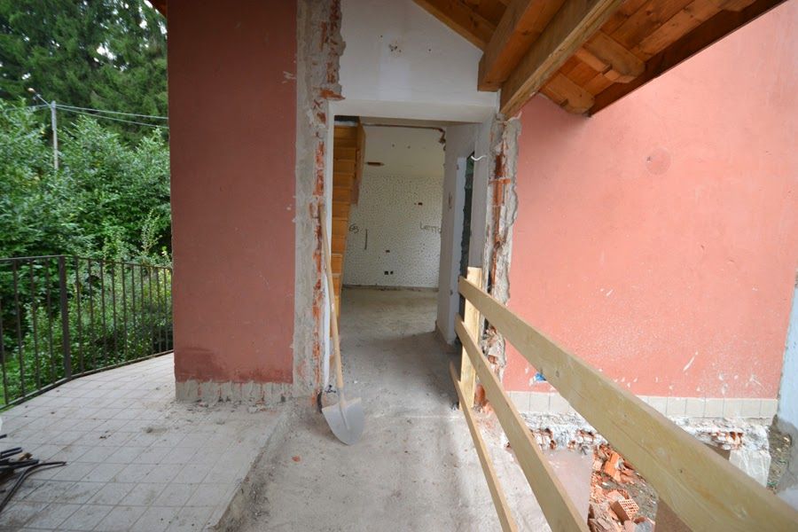 Passaggio coperto parzialmente ristrutturato con pareti rosa, cemento a vista e ringhiera in legno che si affaccia sul verde.
