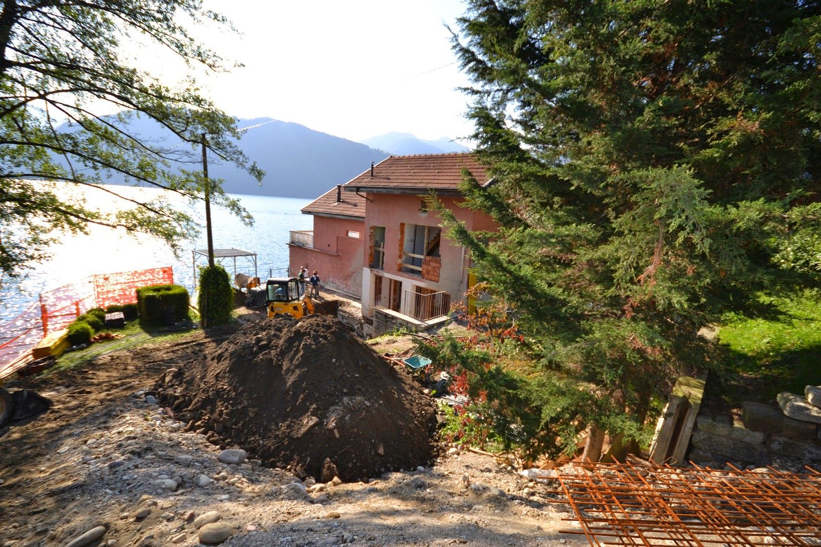 Casa in costruzione sul lago, con cumulo di terra e alberi, affacciata su una vista panoramica di montagne e acqua.