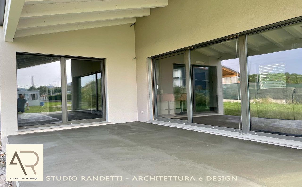 Patio coperto con ampie porte scorrevoli in vetro e travi a vista bianche sul soffitto, affacciato su un paesaggio erboso.