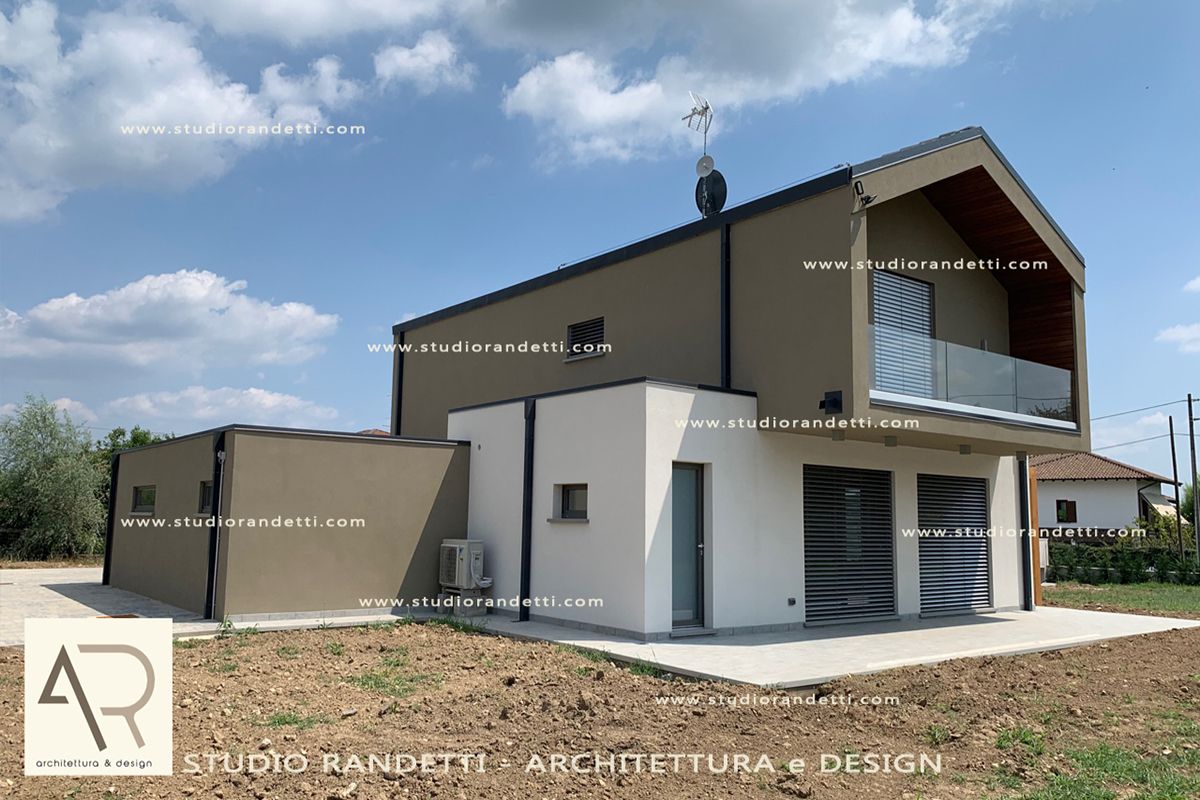 Casa moderna beige a due piani con tetto spiovente, balcone e garage bianco su un lotto erboso sotto un cielo nuvoloso