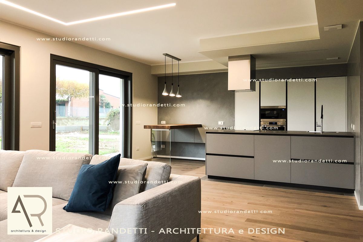 Soggiorno e cucina moderni a pianta aperta con divani grigi, isola elegante, finestre con infissi neri e illuminazione calda.
