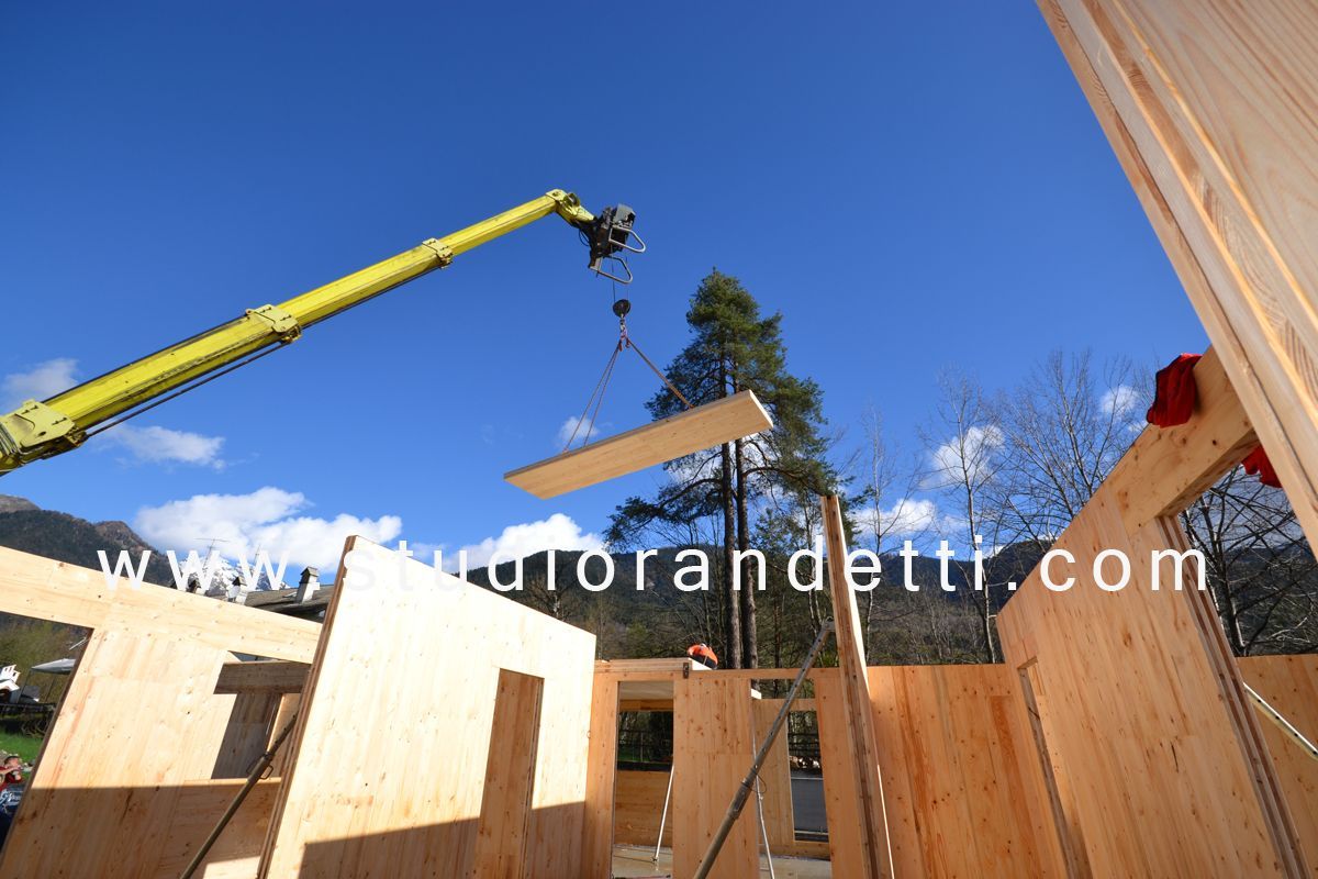 Cantiere edile con gru che solleva un pannello di legno sopra pareti di legno non rifinite sotto un cielo azzurro.