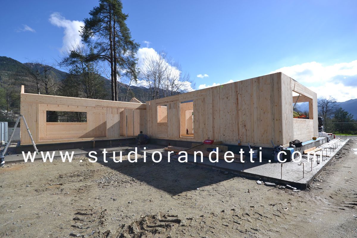 Casa moderna in legno parzialmente costruita su un terreno sterrato con montagne e alberi sullo sfondo.