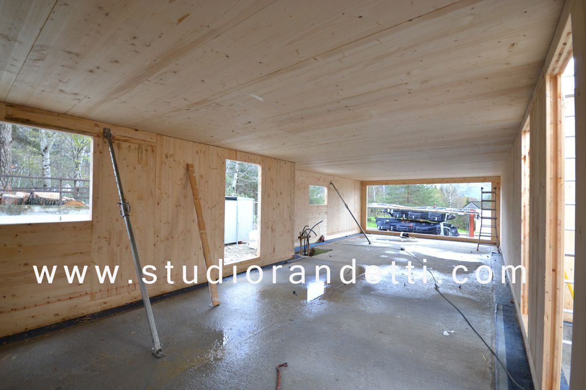 Interno in costruzione con struttura in legno, ampie finestre e pavimento in cemento.