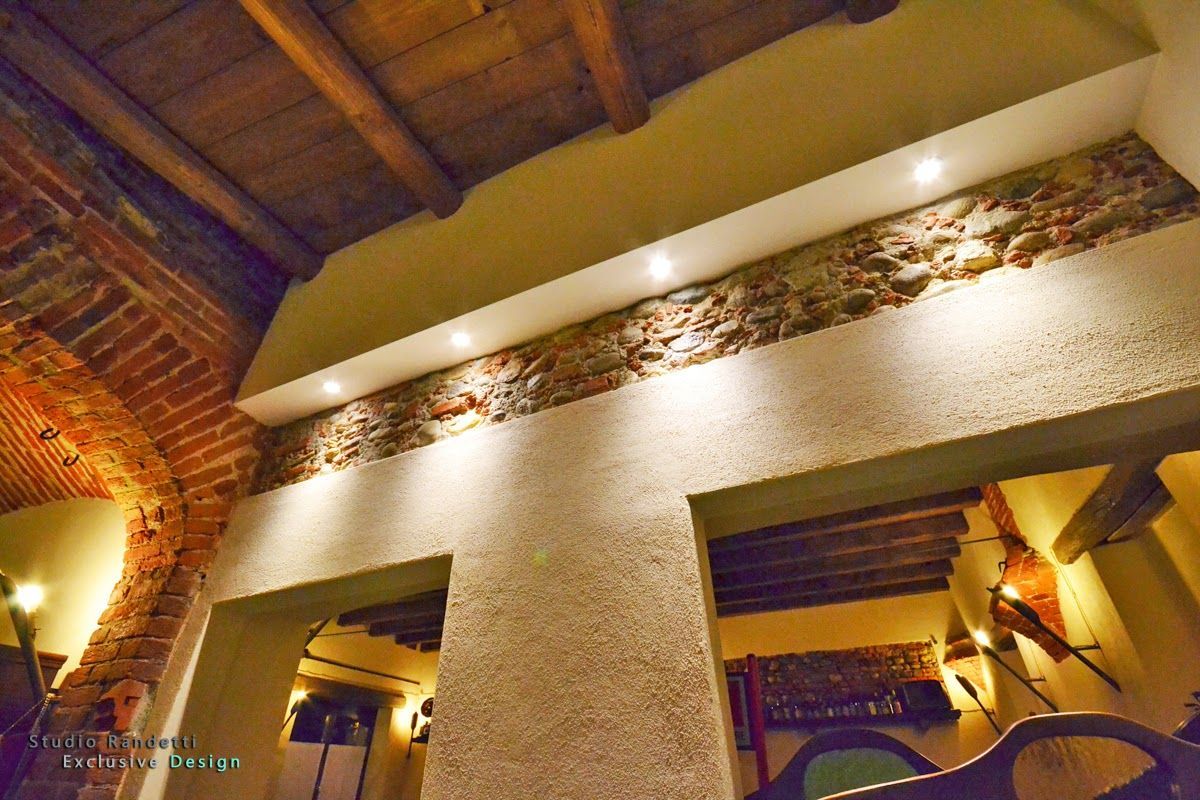 Interno rustico e caldo, illuminato da luci soffuse, con archi in mattoni, pareti con texture e travi in legno sul soffitto.