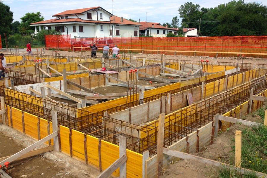 Cantiere edile con casseforme gialle e operai davanti a una casa in costruzione con il tetto rosso.