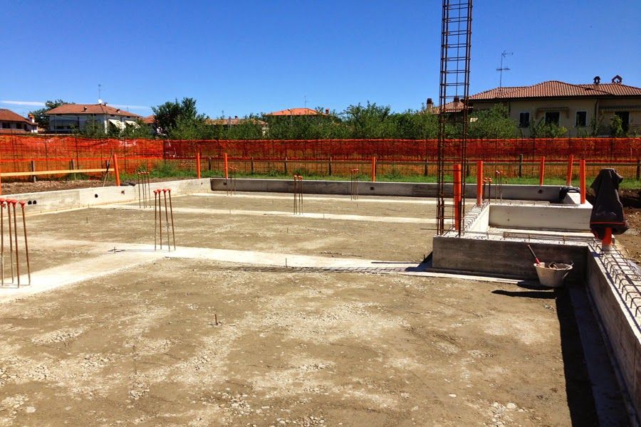 Cantiere edile con fondamenta in cemento, barre d'armatura, recinzione arancione e un operaio vicino al bordo destro.