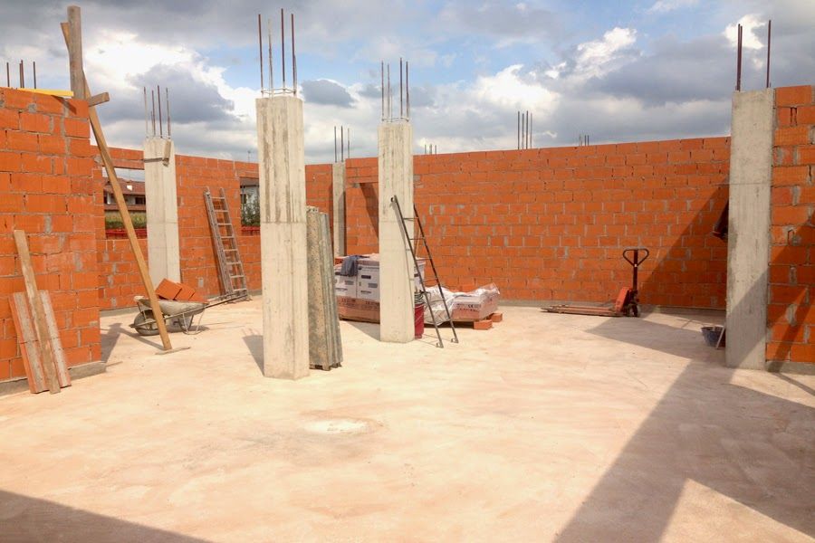 Interno di una casa in mattoni parzialmente costruita, con pavimento in cemento, colonne portanti e materiali edili.