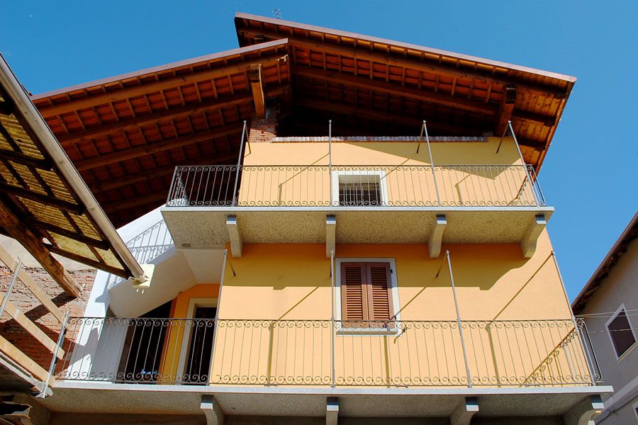 Casa gialla a due piani con balconi e tetto in legno, sullo sfondo di un cielo azzurro.