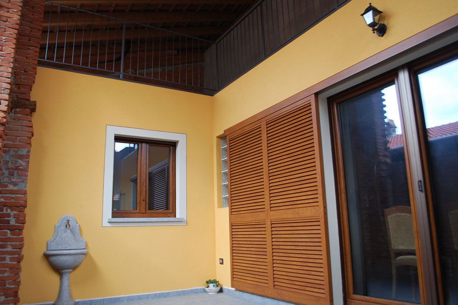 Parete del patio gialla con finestra, porte a persiana in legno e porta scorrevole in vetro sotto un soffitto coperto.