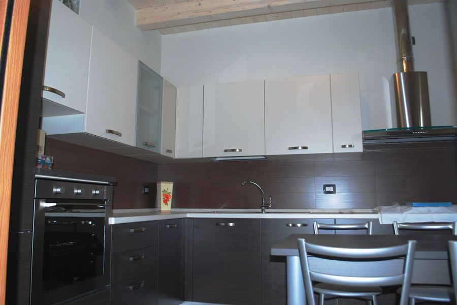 Cucina moderna con mobili bianchi, ripiani scuri, forno in acciaio inox e un piccolo tavolo da pranzo con sedie.