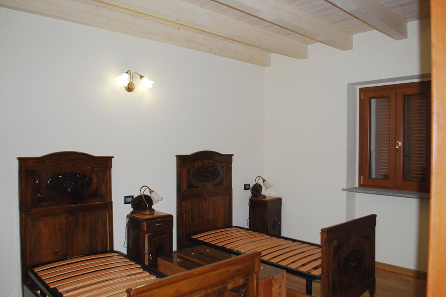 Due letti singoli in legno e comodini in una camera da letto semplice con pareti bianche e travi a vista sul soffitto.