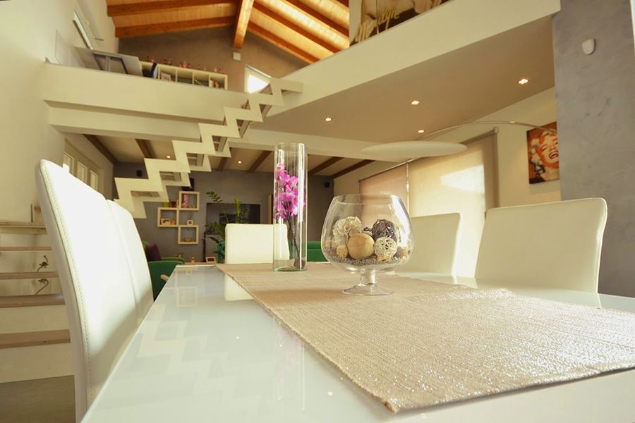 Zona pranzo moderna con sedie bianche, tavolo in vetro, scala e soffitto in legno caldo in un interno luminoso e aperto.